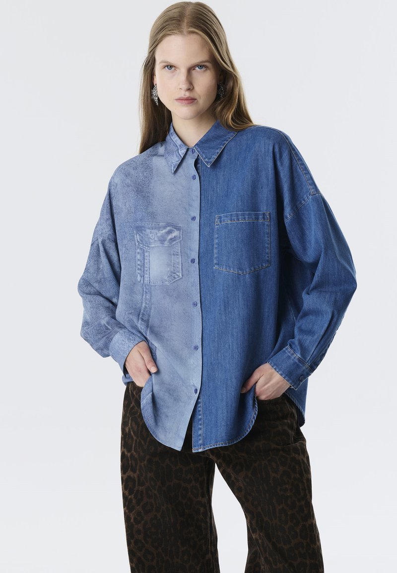 Camisa de mezclilla con un diseño bicolor: un lado azul claro con textura, el otro lado azul oscuro. Mangas largas, cierre con botones.