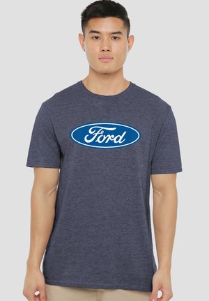 Young man facing forward wearing a navy blue Ford logo t-shirt and beige pants against a plain gray background.