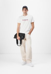 Camiseta blanca con el logo "tommy" en rojo y azul, pantalones beiges, bolsa negra con correa con logo y zapatillas blancas. Una gorra negra completa el look.