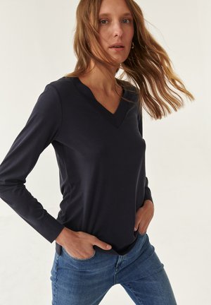 Haut à manches longues, de couleur bleu marine profond, en V, fabriqué dans un tissu lisse, associé à un jean bleu clair. Présente une coupe décontractée et une texture subtile.