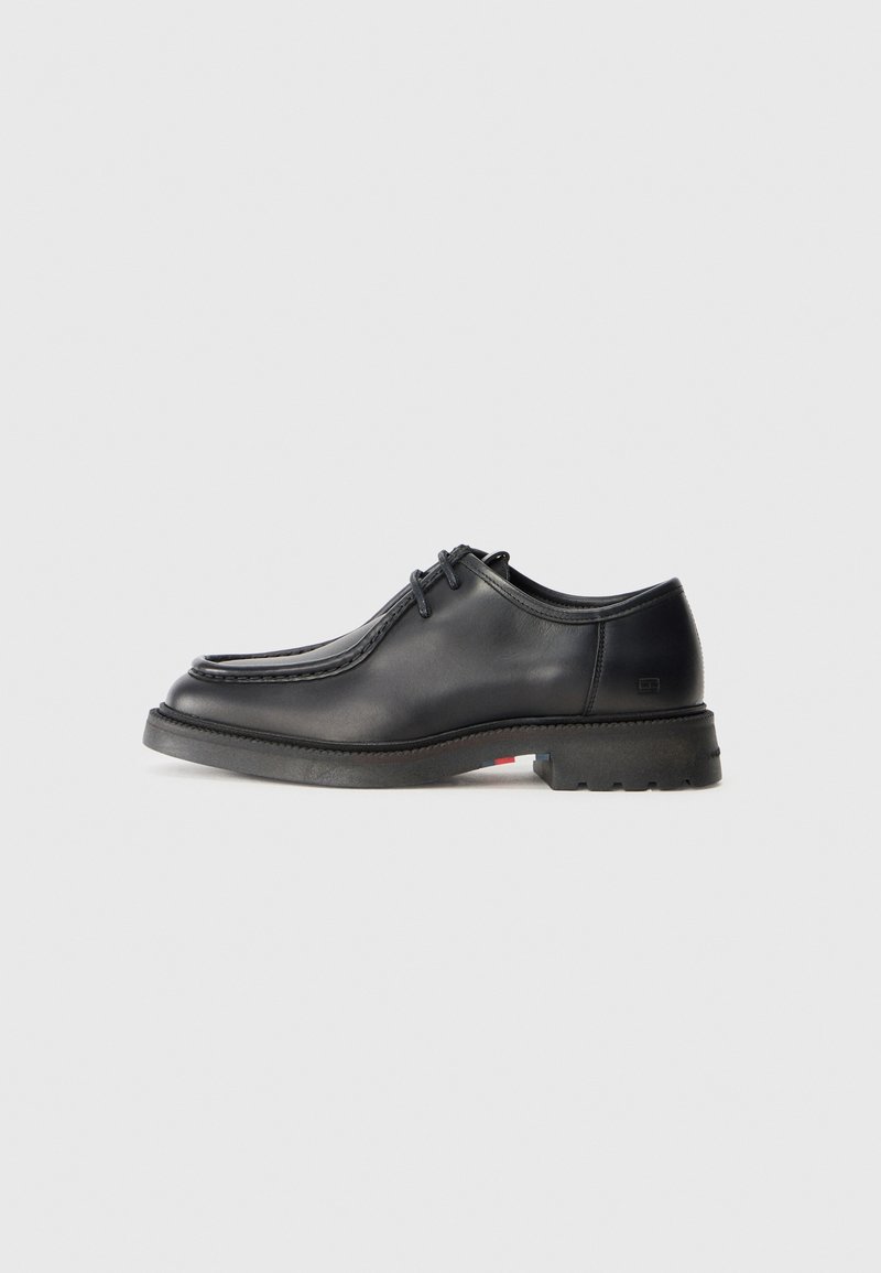 Zapato de cuero negro con textura suave, punta redonda y costura grabada poco profunda. Suela de goma gruesa con un ligero tacón y patrón de agarre.