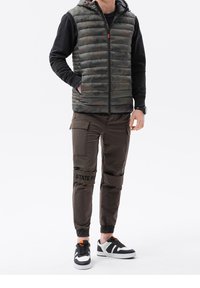 Tarnfarbene Pufferweste mit Kapuze, schwarzes Langarmshirt, braune Cargo-Hosen mit Knieakzenten und schwarze Turnschuhe mit wei�ßen und orangen Details.