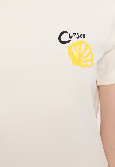 Cremfarbendes kurzärmliges Hemd mit einem schwarz-gelben gestickten Muschel-Design und dem Wort "CLOSEO". Glatte Textur, einfacher Rundhalsausschnitt.
