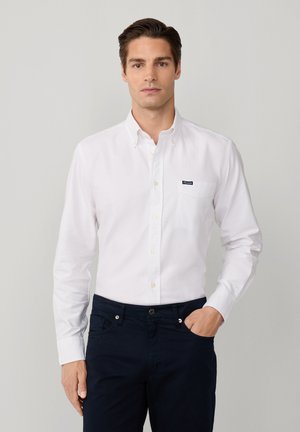 Camicetta bianca con colletto, dotata di taschino e piccolo logo. Indossata con pantaloni blu scuro. Il tessuto appare liscio, leggero e privo di texture.
