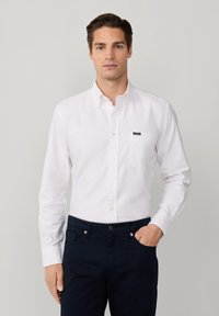 Camicetta bianca con colletto, dotata di taschino e piccolo logo. Indossata con pantaloni blu scuro. Il tessuto appare liscio, leggero e privo di texture.