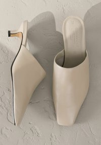 Beige nahka-muleja, joissa on terävä kärki, avoin takaosa ja korkea korko, jota korostaa musta yksityiskohta kantapäässä. Sujuva rakenne ja minimalistinen muotoilu.