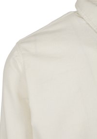 Gros plan sur l’épaule et le col d’une chemise en tissu côtelé de couleur crème, montrant la texture et les détails des coutures.