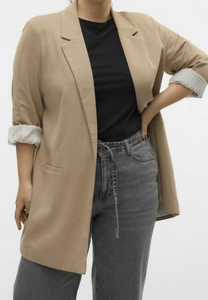 Kvinde iført beige blazer med op rullede ærmer, sort skjorte og grå høj taljede jeans, stående med den ene hånd på hoften.