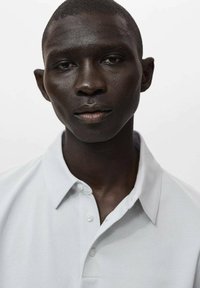 Vit polo skjorta med krage, med två knappar vid halsringningen. Slät textur, minimalistisk design och inga synliga mönster.