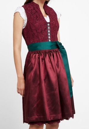 Kvinde iført en vinrød blonde dirndl-kjole med sølvknapper, hvid blondebluse og et grønt satinbånd bundet om livet.