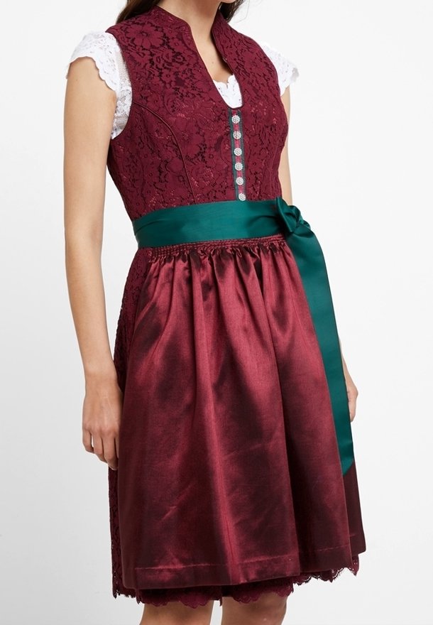 Femme portant une robe dirndl en dentelle bordeaux avec des boutons argentés, un chemisier en dentelle blanc et un ruban de satin vert noué à la taille.