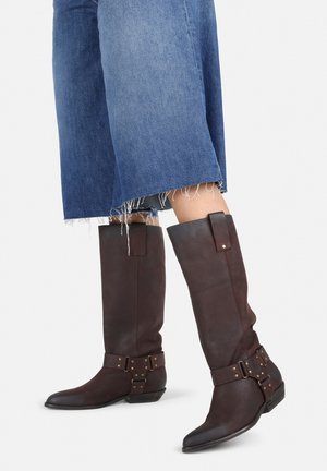 Botas de cuero marrón hasta la rodilla con un diseño ajustado, hebillas decorativas y una puntera ligeramente puntiaguda. Usadas con culottes de mezclilla con dobladillo deshilachado.