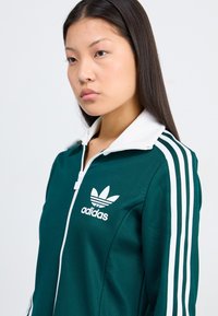 En ung kvinna med långt, rakt hår bär en grön zip-up-jacka med vita ränder och en Adidas-logotyp, vänd något åt sidan.