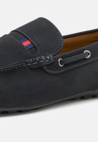 Des mocassins en daim bleu marine foncé présentent un accent cousu avec des rayures rouges et bleues, des œillets métalliques et une semelle texturée pour une meilleure adhérence.