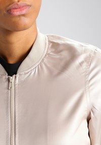 Světlý béžový bomber s hladkou, lesklou látkou, žebrovaným límcem a předním zipem. Jednoduchý design bez vzorů nebo kovových doplňků.