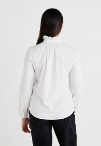White long-sleeve shirt with a high ruffled collar, gathered back, and fitted sleeves, paired with dark jeans. Smooth texture.