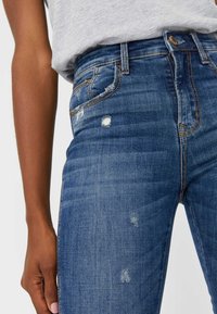 Persona vistiendo un pantalón de mezclilla azul desgastado y una camisa clara gris metida por dentro, con la mano descansando al lado de la pierna.