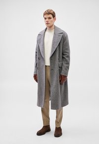 Μακρύ γκρι παλτό ψαροκόκαλο με φαρδιές πέτσες, σχεδιασμένο διπλής όψης, σε συνδυασμό με εκρού ριμπ πλεκτό πουλόβερ, μπεζ παντελόνια και καφέ γάντια.