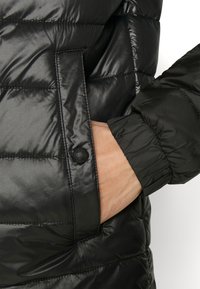 Giacca a vento nera con finitura lucida, design trapuntato, polsini elasticizzati e tasca con bottone, che mette in mostra uno stile elegante e funzionale.