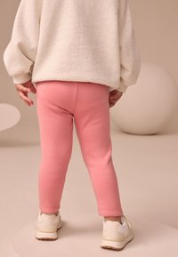 Colanți roz deschis din material moale, cu croială mulată, purtați cu un pulover supradimensionat de culoare crem și adidași albi.