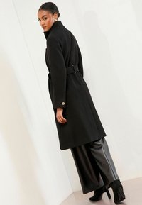 Melna vilnas mēteli ar augstu apkakli, jostasvietu un pogātām piedurknēm, apvienots ar spīdīgām melnām platām biksēm un ar papēžiem apaviem līdz potītēm.