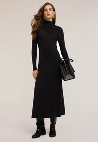 Abito nero a maniche lunghe con collo alto, texture a costine, vestibilità slim e gonna svasata. Abbinato a una borsa nera e stivali fino al ginocchio.