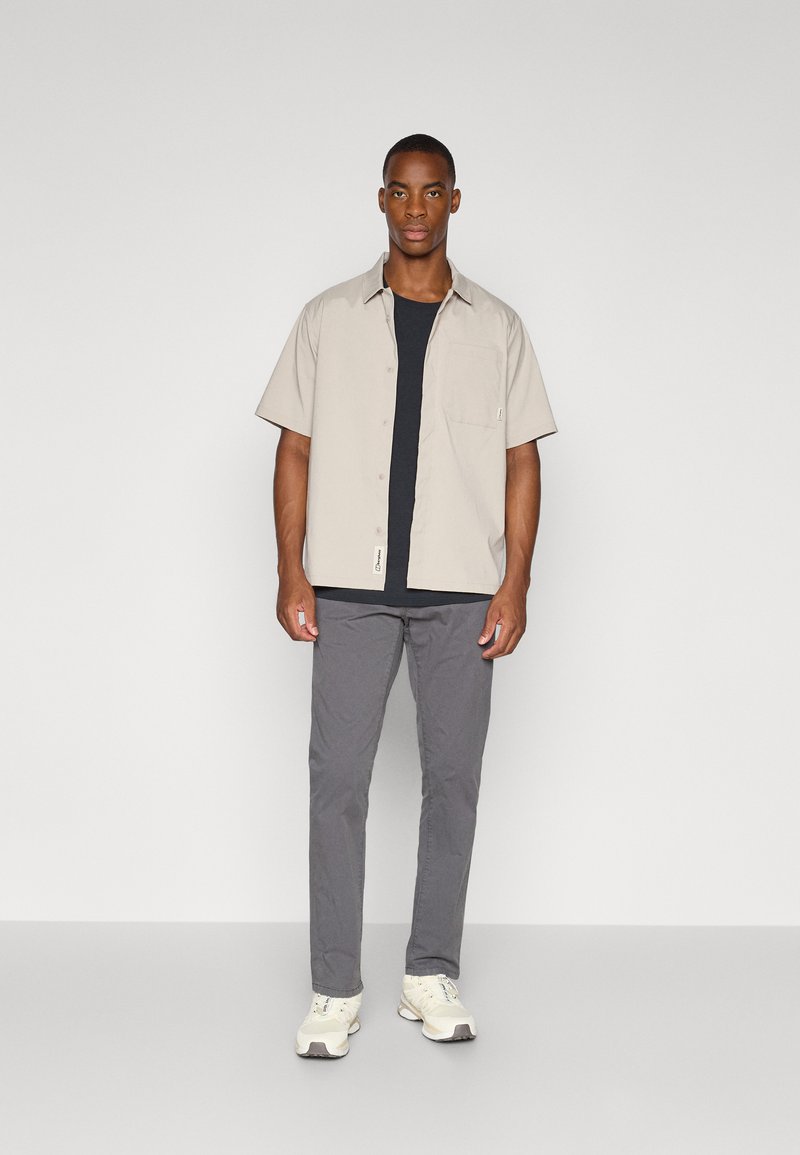 Lässiges Outfit mit einem hellbeigen Kurzarmhemd mit Tasche über einem schwarzen T-Shirt, kombiniert mit grauen Hosen und weißen Sneakers.