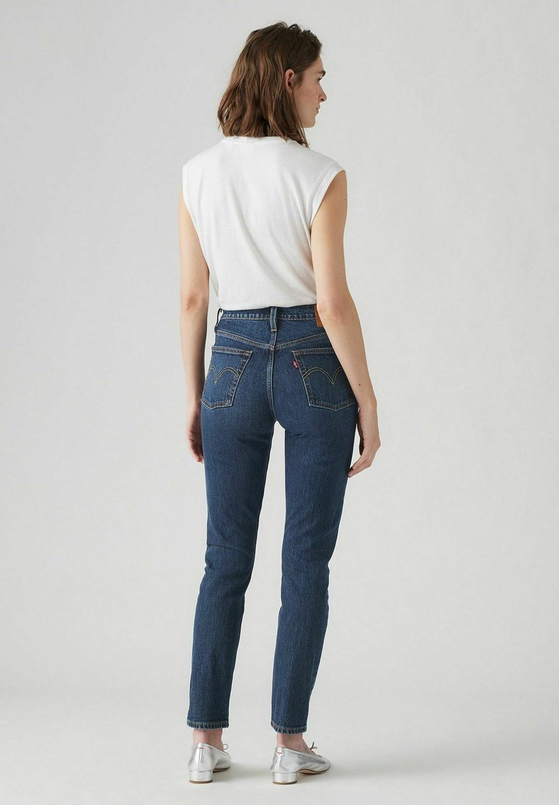 Jean en denim bleu foncé taille haute avec une coupe slim, présentant des poches arrière avec des coutures jaunes et une étiquette Levi's sur la ceinture.