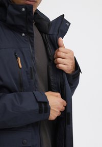 Tamsiai mėlyna striukė su užtrauktuku, spaudėmis ir velcro akcentais. Turinti išklotą apykaklę ir kelias kišenes, pagaminta iš tvirtos medžiagos.
