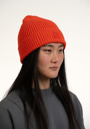 Orange knit beanie with vertical ribbing, featuring a rectangular logo patch. Smooth texture, tapered top, and fitted design.