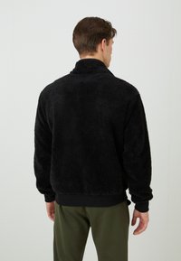 Sort fleecejakke med høj krave, blød tekstur og ribbede manchetter. Ryggen har en løs pasform, og der er ingen synlige logoer eller accenter.