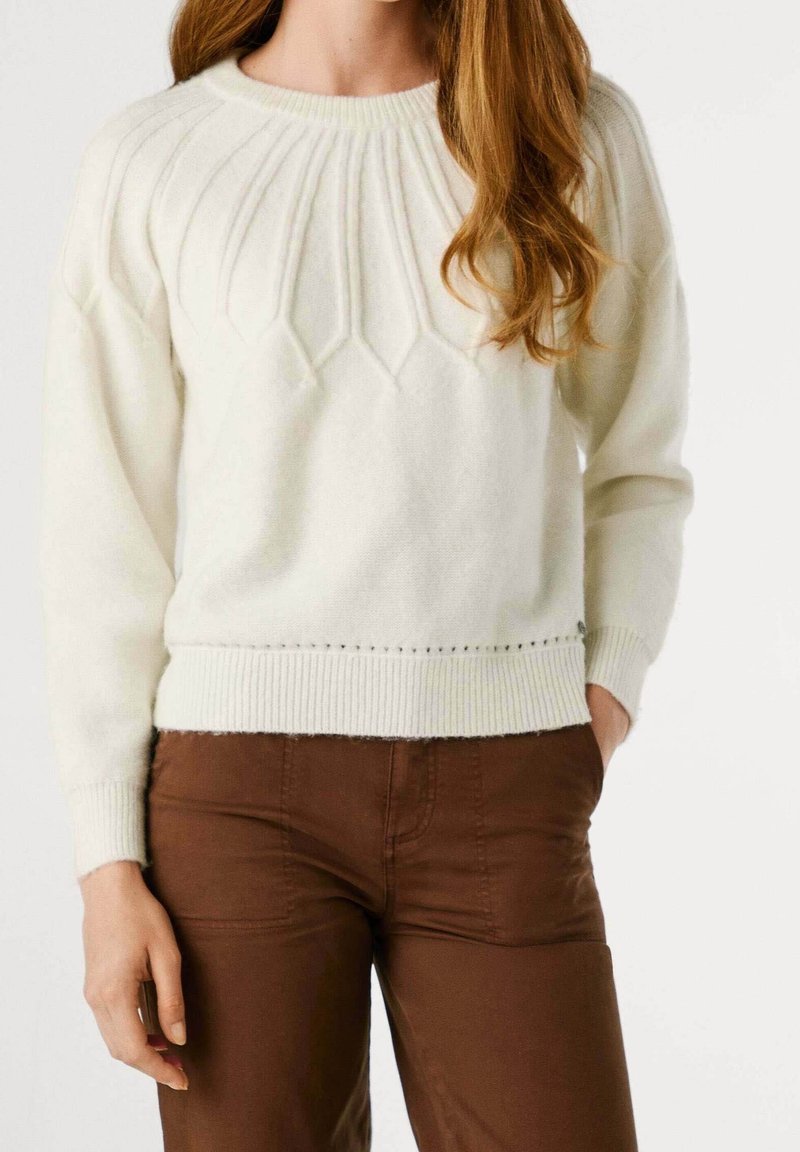 Femme portant un pull en maille crème avec un détail texturé sur le yoke, associé à un pantalon marron taille haute, les mains détendues le long du corps.