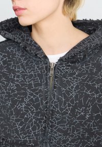 Black zip-up jacket with a textured cracked pattern, featuring a hood and frayed edges along the collar, made from a soft fabric.