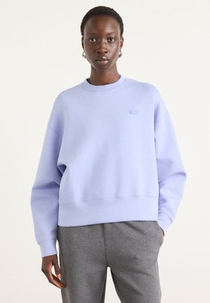 Sweat-shirt lilas clair avec un col rond, des épaules tombantes et des poignets côtelés. Il arbore un petit logo brodé sur la poitrine.