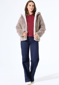Mujer vestida con una chaqueta de piel sintética beige sobre un suéter burdeos, jeans azules y zapatillas blancas, de pie frente a un fondo blanco liso.