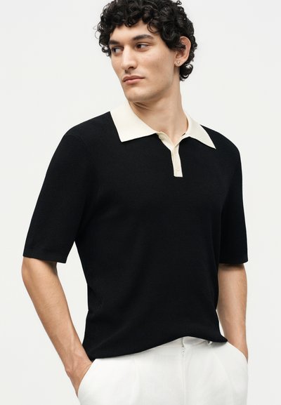 Νέος άνδρας με σγουρά μαλλιά που φοράει μαύρο πόλο με κοντά μανίκια και λευκό γιακά, λευκό παντελόνι, με τα χέρια στις τσέπες, κοιτάζει πλαγίως.