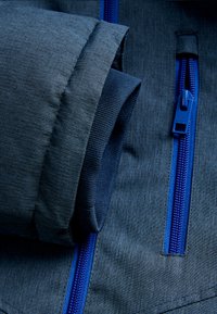 Veste bleu marine en tissu lisse et texturé. Présente des zips et des passepoils bleus contrastants. Extrémités de manches roulées visibles.