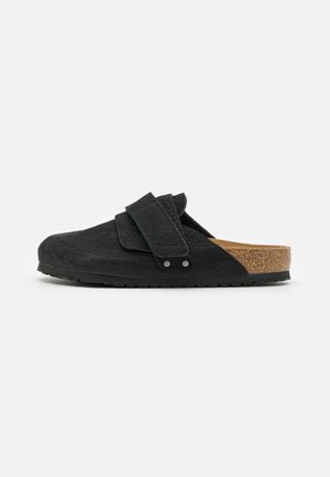 Black slip-on clog made of textured material with a cork footbed, featuring an adjustable strap and rubber outsole.