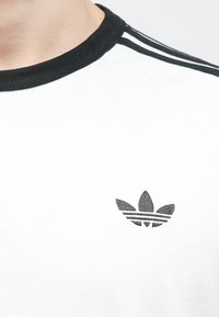 Biela tričko s čiernym rebrovaným golierom, s tromi čiernymi pruhmi pozdĺž ramien a malým čiernym vyšitým logom.