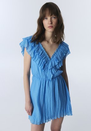 Jeune femme portant une robe bleu clair plissée à volants avec un décolleté en V profond, debout devant un fond clair uni.
