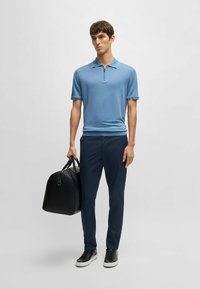 Light blue polo shirt with a zippered neck, paired with dark navy trousers and black sneakers. Carrying a black leather duffel bag.
