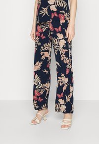 Blommiga marinblå byxor med rosa och krämfärgade botaniska mönster, vida ben, lätt material, kombinerade med vita sandaletter med klack.