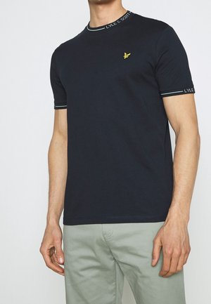 Mężczyzna w dopasowanej czarnej koszulce Lyle & Scott z logo, z markowanym kołnierzem i krawędziami rękawów, w połączeniu z jasnoszarymi spodniami.