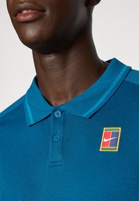 Tyrkysové polo tričko Nike s textúrovanou látkou, obsahujúce trojité zapínanie a nášivku loga v červenej, žltej a modrej farbe na ľavej strane hrudi.