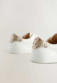 Baskets en cuir blanc avec une surface texturée, présentant un accent pailleté doré et argenté sur le talon et une doublure intérieure beige.
