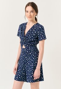 Jumpsuit blu navy con motivo floreale, maniche corte, dettaglio anteriore annodato, scollatura in vita e pantaloni corti dal taglio rilassato.