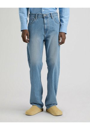 Mężczyzna ubrany w jasnoniebieskie dżinsy, jasnoniebieską koszulę na guziki oraz beżowe wsuwane buty stoi na jednolitym tle.