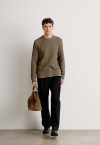 Olivgrüner strukturierter Strickpullover, schwarze Hose und schwarze Schuhe. Hält eine braune Lederhandtasche. Schlichte, zeitgenössische Silhouette.