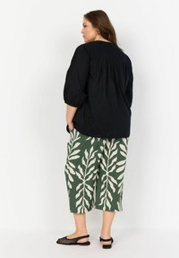 Blusa nera con maniche a sbuffo, vestibilità rilassata. Culotte verdi con motivo a foglia bianca. Scarpe slip-on nere in rete, con tasche laterali visibili.