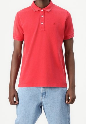 Homme portant un polo rouge à manches courtes avec trois boutons et un jean en denim bleu clair, debout devant un fond uni.